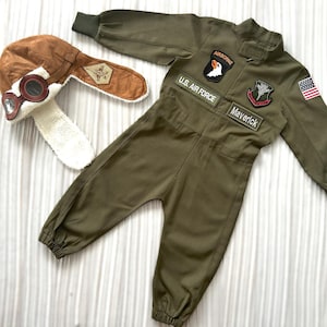 Könnte beinhalten: Ein grüner Overall mit Aufnähern, auf denen "Airborne", "U.S. Air Force", "Maverick" und eine kleine amerikanische Flagge stehen. Der Overall hat einen Reißverschluss und elastische Bündchen. Eine braune Fliegermütze mit Kunstfellbesatz und einer Brille ist im Lieferumfang enthalten.