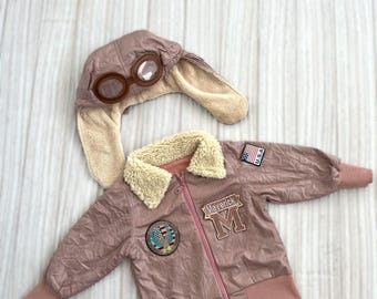 Könnte beinhalten: Eine staubrosafarbene Fliegerjacke und ein passender Hut mit Brille. Die Jacke hat einen cremefarbenen Kunstfellkragen, einen Reißverschluss und Aufnäher mit der Aufschrift "Maverick" und "U.S.A.". Der Hut hat Ohrenklappen und eine klare Brille.