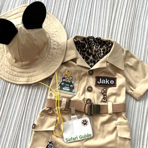 May include: A tan safari outfit for a child with a safari guide badge, a zebra patch, a lion patch, and a jeep patch. The outfit includes a hat with Mickey Mouse ears, a short-sleeved shirt, and shorts with a belt. The name "Jake" is written on a name tag.