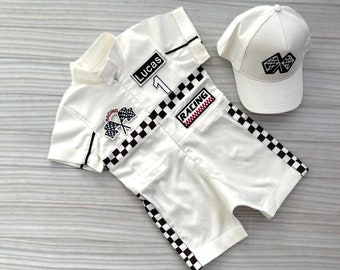 Op de afbeelding: Wit racepak met zwart-witte ruitjesbies. Het pak heeft het nummer "1" en het woord "Lucas" op de voorkant. Het heeft ook een patch met de tekst "Racing" en een patch met gekruiste ruitjesvlaggen. Een witte baseballpet met ruitjesvlaggen op de voorkant.