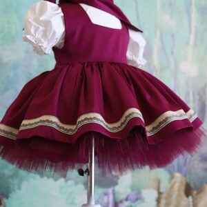 Peut inclure: Une robe bordeaux avec un col blanc et une manche bouffante blanche. La robe a une jupe en tulle et une bordure d&eacute;corative autour de l'ourlet.
