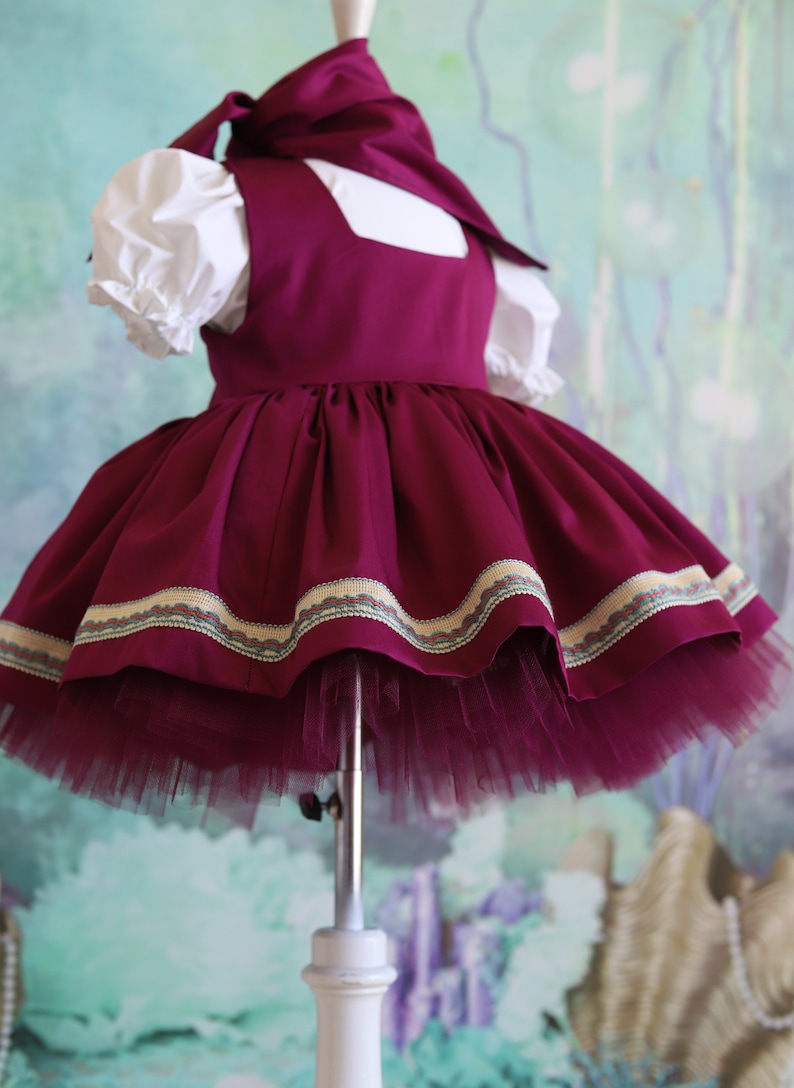 Peut inclure: Une robe bordeaux avec un col blanc et une manche bouffante blanche. La robe a une jupe en tulle et une bordure d&eacute;corative autour de l'ourlet.