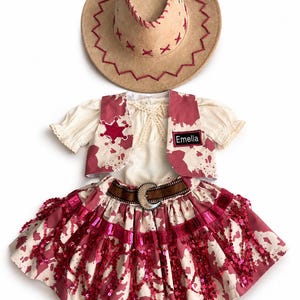 Può includere: Un cappello da cowboy beige con cuciture rosse e un completo abbinato. Il completo include un top color crema, un gilet con una stella, una gonna con una cintura marrone e il nome "Emella". La gonna ha paillettes rosse.