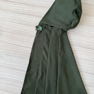 May include: A dark green cloak with an attached hood. The cloak is secured with a decorative leaf-shaped clasp. The fabric appears to be a soft, flowing material. The cloak is designed to be worn over the shoulders.