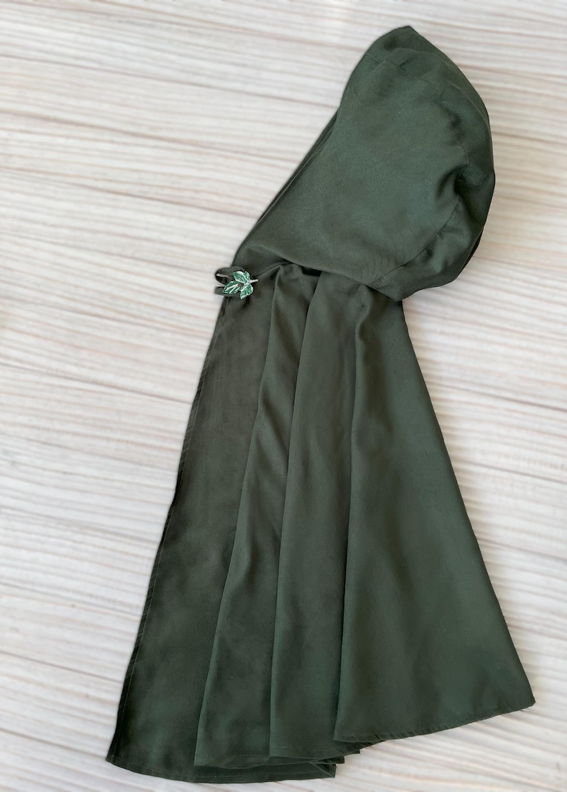 May include: A dark green cloak with an attached hood. The cloak is secured with a decorative leaf-shaped clasp. The fabric appears to be a soft, flowing material. The cloak is designed to be worn over the shoulders.