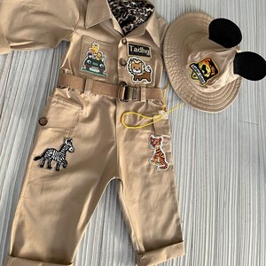 May include: A tan safari jumpsuit with a yellow belt and several animal patches. The jumpsuit has a zipper closure and two pockets. The jumpsuit is paired with a tan safari hat with black Mickey Mouse ears.