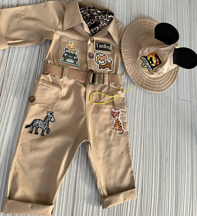 May include: A tan safari jumpsuit with a yellow belt and several animal patches. The jumpsuit has a zipper closure and two pockets. The jumpsuit is paired with a tan safari hat with black Mickey Mouse ears.
