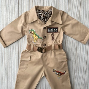 Pu&ograve; includere: Una tuta safari beige con una cintura marrone e toppe di dinosauri. La tuta ha una fodera con stampa leopardata e un'etichetta con il nome "Kailee 1".
