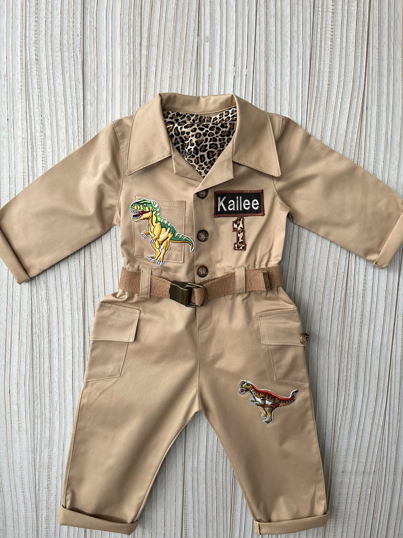 Pu&ograve; includere: Una tuta safari beige con una cintura marrone e toppe di dinosauri. La tuta ha una fodera con stampa leopardata e un'etichetta con il nome "Kailee 1".