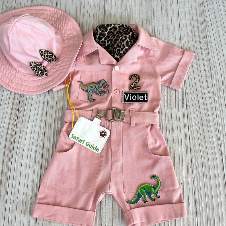 May include: Pink safari jumpsuit with a dinosaur patch, a leopard print fabric lining, and a name tag that says "Violet".