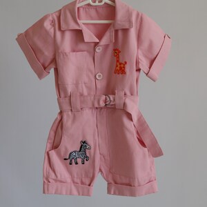 K&ouml;nnte beinhalten: Ein rosa Baby-Overall mit kurzen &Auml;rmeln und einem G&uuml;rtel. Der Overall hat eine Giraffe und ein Zebra, die auf der Vorderseite gestickt sind.