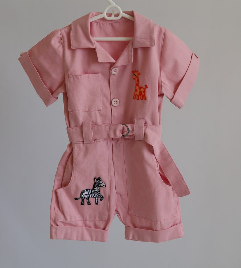 K&ouml;nnte beinhalten: Ein rosa Baby-Overall mit kurzen &Auml;rmeln und einem G&uuml;rtel. Der Overall hat eine Giraffe und ein Zebra, die auf der Vorderseite gestickt sind.