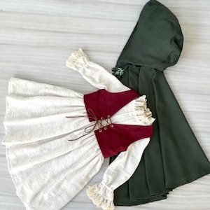 May include: A cream-colored dress with long sleeves and a burgundy vest with a lace-up front. The sleeves have lace cuffs. A dark green hooded cloak is next to the dress. The outfit is likely for a child.