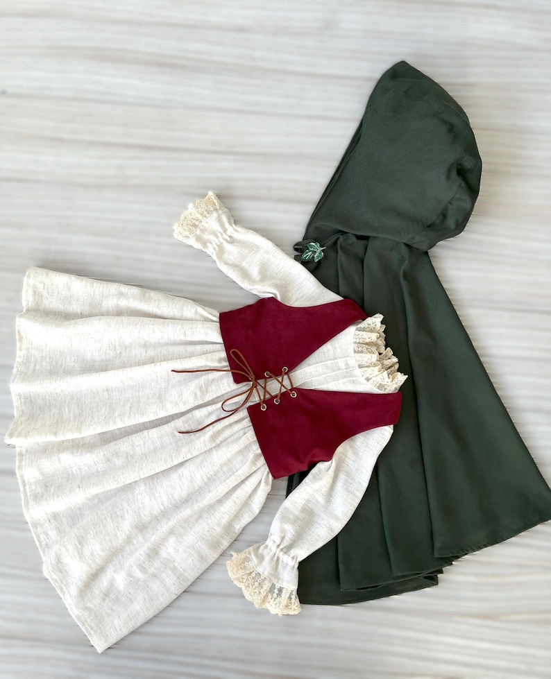 May include: A cream-colored dress with long sleeves and a burgundy vest with a lace-up front. The sleeves have lace cuffs. A dark green hooded cloak is next to the dress. The outfit is likely for a child.