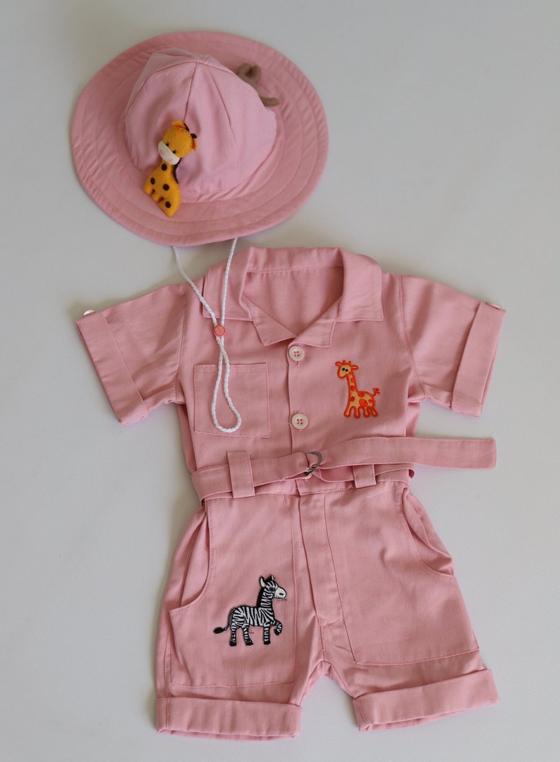 K&ouml;nnte beinhalten: Rosafarbenes Safari-Outfit f&uuml;r Babys mit passendem Hut. Das Outfit besteht aus einem kurz&auml;rmeligen Overall mit Kn&ouml;pfen vorne, einem G&uuml;rtel und gestickten Bildern einer Giraffe und eines Zebras. Der Hut ist ein breitkrempiger Sonnenhut mit einer gestickten Giraffe.