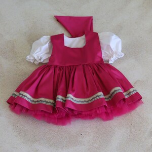 K&ouml;nnte beinhalten: Ein rosa Kleid mit einem wei&szlig;en Oberteil und einem T&uuml;llrock. Das Kleid hat einen dekorativen Besatz am Saum und ein passendes rosa Kopftuch.
