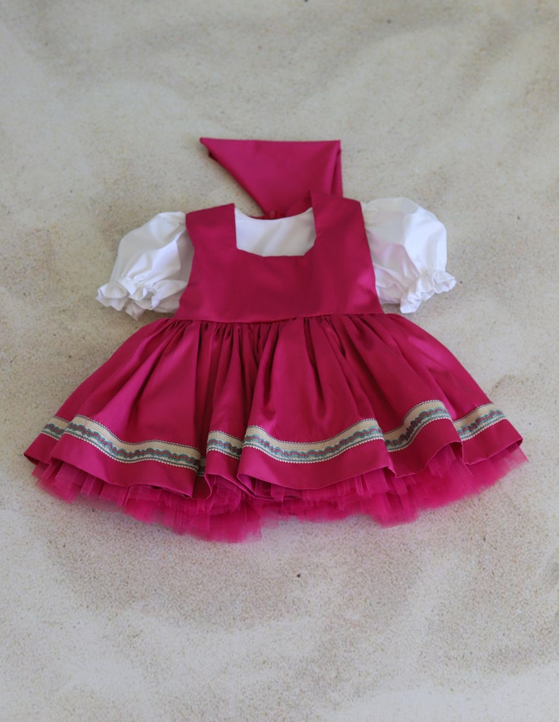 K&ouml;nnte beinhalten: Ein rosa Kleid mit einem wei&szlig;en Oberteil und einem T&uuml;llrock. Das Kleid hat einen dekorativen Besatz am Saum und ein passendes rosa Kopftuch.