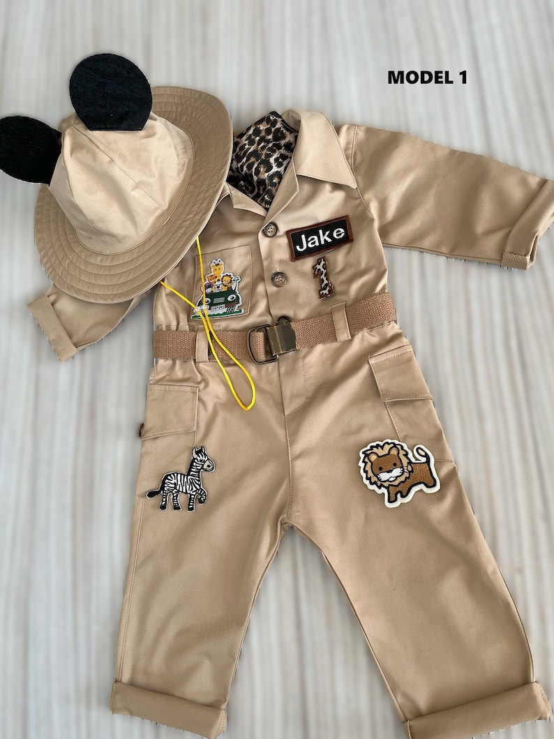 May include: A tan safari jumpsuit with a black and white zebra, a brown lion, and a green jeep patch. The jumpsuit has a brown belt with a yellow string attached. The outfit also includes a tan hat with black Mickey Mouse ears and a brown and black leopard print scarf.