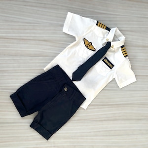 May include: A white pilot uniform set with a black tie and shorts. The shirt has gold epaulettes, a pilot's wings patch, and a name tag that reads "Jaxson". The shorts are navy blue.