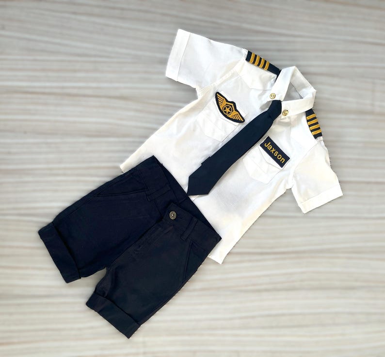 May include: A white pilot uniform set with a black tie and shorts. The shirt has gold epaulettes, a pilot's wings patch, and a name tag that reads "Jaxson". The shorts are navy blue.