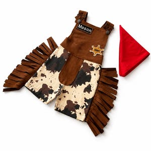 Op de afbeelding: Een bruin en wit cowboy-outfit met franjes, een sheriff-badge en een rode bandana. De outfit bevat een tuinbroek met de naam "Mason". De tuinbroek heeft een koeienhuidpatroon.