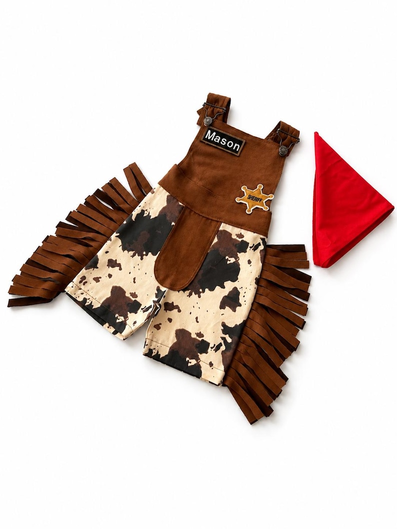 Op de afbeelding: Een bruin en wit cowboy-outfit met franjes, een sheriff-badge en een rode bandana. De outfit bevat een tuinbroek met de naam "Mason". De tuinbroek heeft een koeienhuidpatroon.