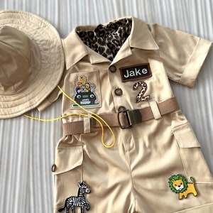 May include: A tan safari-themed outfit for a child, featuring a short-sleeved jumpsuit with a belt, a safari hat, and various animal patches. The jumpsuit has a pocket with a jeep and animal patch, a name tag that says "Jake", and a number "2" patch. The outfit also has a zebra and lion patch.