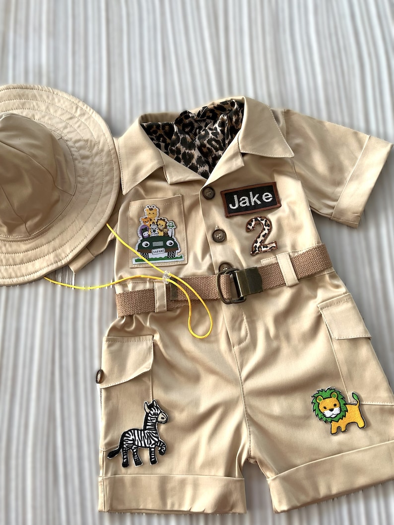 May include: A tan safari-themed outfit for a child, featuring a short-sleeved jumpsuit with a belt, a safari hat, and various animal patches. The jumpsuit has a pocket with a jeep and animal patch, a name tag that says "Jake", and a number "2" patch. The outfit also has a zebra and lion patch.