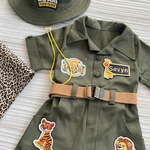 May include: A green safari jumpsuit with a brown belt and several patches, including a tiger, a lion, a giraffe, and a safari tour guide patch. The jumpsuit has a yellow lanyard and a name tag that says "Sevyyn".