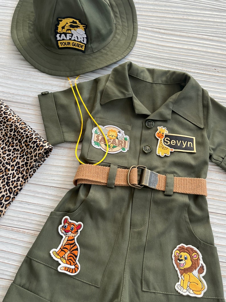 May include: A green safari jumpsuit with a brown belt and several patches, including a tiger, a lion, a giraffe, and a safari tour guide patch. The jumpsuit has a yellow lanyard and a name tag that says "Sevyyn".