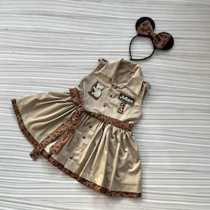 May include: A tan dress with a brown leopard print trim and belt, featuring a koala patch and the name "Julian". A matching headband with mouse ears and a leopard print bow is also included. The dress has a button-down front.