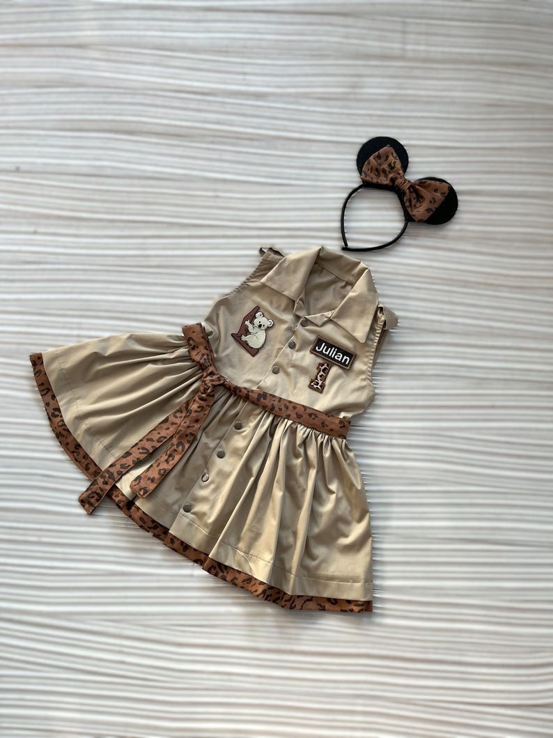 May include: A tan dress with a brown leopard print trim and belt, featuring a koala patch and the name "Julian". A matching headband with mouse ears and a leopard print bow is also included. The dress has a button-down front.