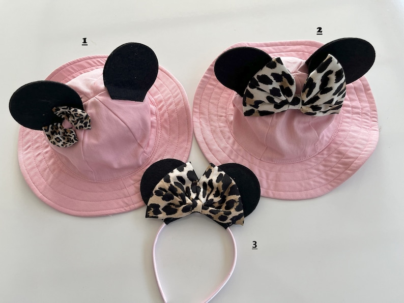 May include: Two pink sun hats with black ears and leopard print bows. A headband with black ears and a leopard print bow.