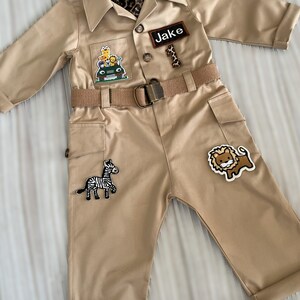 May include: A tan safari jumpsuit with a brown belt and a name tag that says "Jake". The jumpsuit has several patches of animals, including a zebra, a lion, and a giraffe.