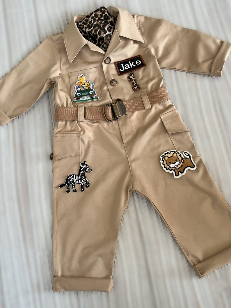 May include: A tan safari jumpsuit with a brown belt and a name tag that says "Jake". The jumpsuit has several patches of animals, including a zebra, a lion, and a giraffe.