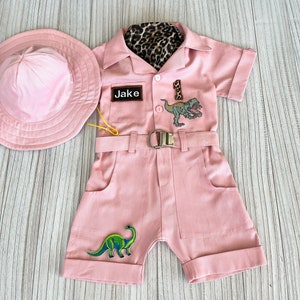 May include: Pink safari jumpsuit with a brown leopard print collar, a name tag that says "Jake", and dinosaur patches. The jumpsuit has a belt and pockets. A pink hat is included.