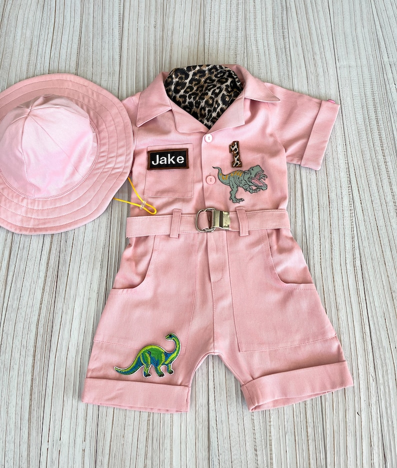 May include: Pink safari jumpsuit with a brown leopard print collar, a name tag that says "Jake", and dinosaur patches. The jumpsuit has a belt and pockets. A pink hat is included.