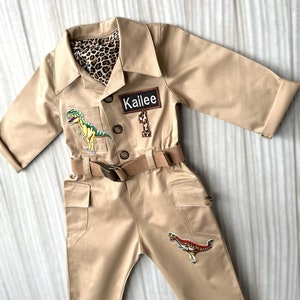 Pu&ograve; includere: Una tuta da safari beige con una cintura marrone e toppe di dinosauri. La tuta ha un'etichetta con il nome "Kailee".