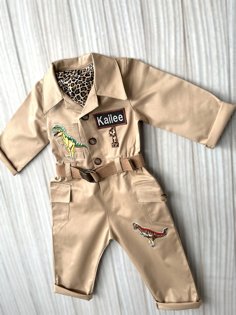 Pu&ograve; includere: Una tuta da safari beige con una cintura marrone e toppe di dinosauri. La tuta ha un'etichetta con il nome "Kailee".
