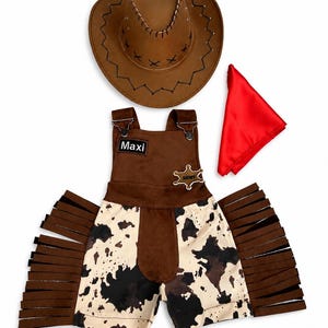 Op de afbeelding: Een bruine cowboyhoed, een rode bandana en een bruin-witte outfit met koeienprint en franjes. De outfit bevat een korte broek en een overall met de naam "Maxi" op een patch en een sheriff-badge.