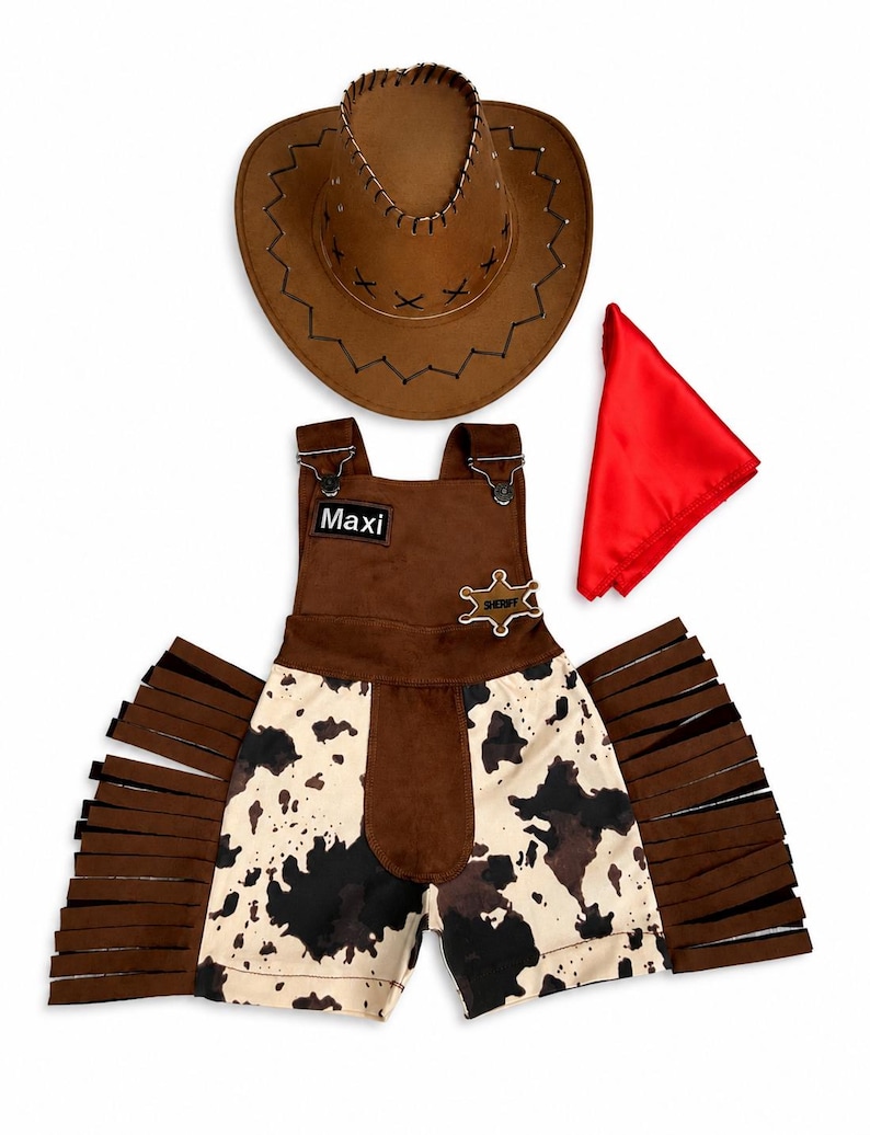 Op de afbeelding: Een bruine cowboyhoed, een rode bandana en een bruin-witte outfit met koeienprint en franjes. De outfit bevat een korte broek en een overall met de naam "Maxi" op een patch en een sheriff-badge.
