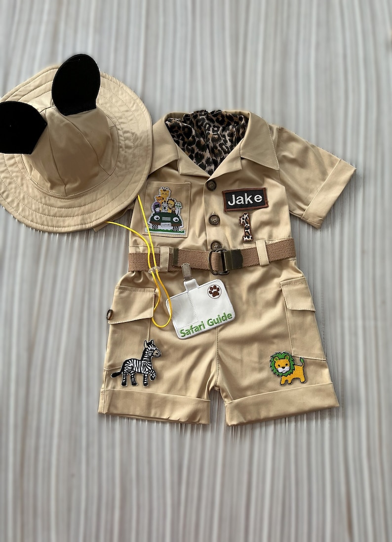 May include: A tan safari-themed outfit for a child, featuring a safari hat with Mickey Mouse ears, a tan jumpsuit with a brown belt, and various patches including a zebra, a lion, and a jeep with animals. The jumpsuit has a pocket with a white tag that says "Safari Guide". The name "Jake" is written on a patch on the chest.