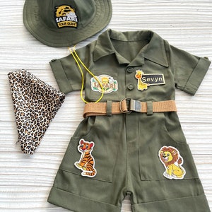 May include: A green safari-themed outfit for a child, featuring a hat with the text "Safari Tour Guide", a short-sleeved jumpsuit with pockets, a belt, and various animal patches. The name "Sevyvn" is written on a patch on the jumpsuit.