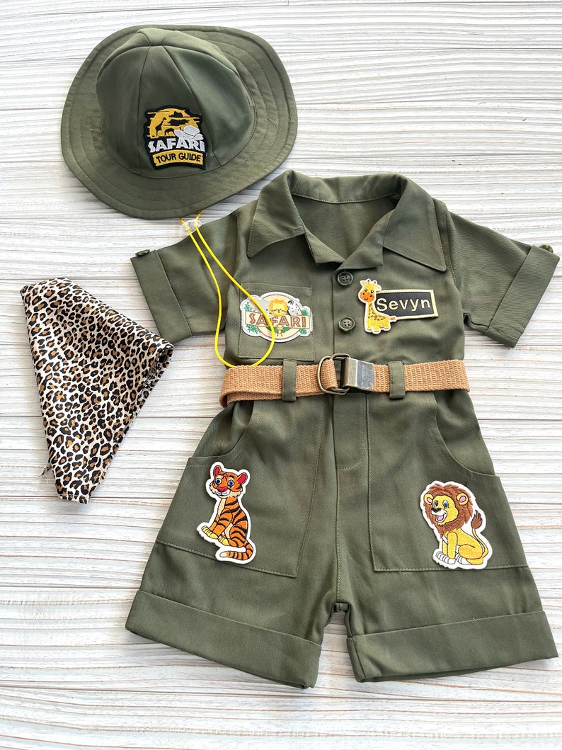 May include: A green safari-themed outfit for a child, featuring a hat with the text "Safari Tour Guide", a short-sleeved jumpsuit with pockets, a belt, and various animal patches. The name "Sevyvn" is written on a patch on the jumpsuit.
