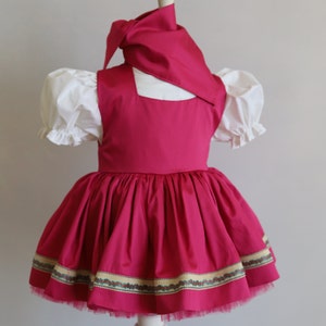K&ouml;nnte beinhalten: Ein rosa Kleid mit einer wei&szlig;en Bluse und einem rosa Kopftuch. Das Kleid hat einen gerafften Rock mit einem gr&uuml;n-goldenen Besatz.