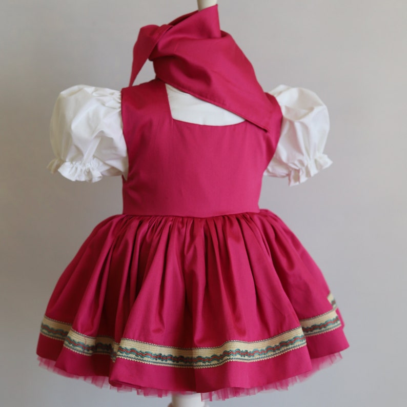 K&ouml;nnte beinhalten: Ein rosa Kleid mit einer wei&szlig;en Bluse und einem rosa Kopftuch. Das Kleid hat einen gerafften Rock mit einem gr&uuml;n-goldenen Besatz.