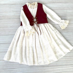 May include: A white dress with long sleeves, a burgundy vest, and a lace-trimmed collar and cuffs. The vest has a brown lace-up detail. The dress has a full skirt and is made of a textured fabric.