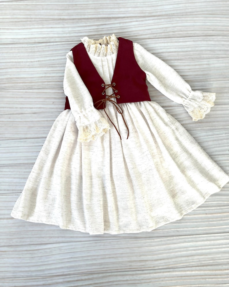 May include: A white dress with long sleeves, a burgundy vest, and a lace-trimmed collar and cuffs. The vest has a brown lace-up detail. The dress has a full skirt and is made of a textured fabric.