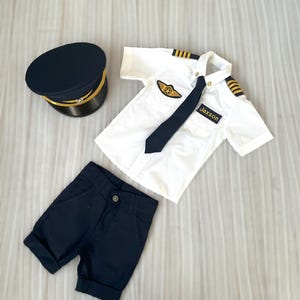 May include: A navy blue pilot's uniform set, including a hat, shirt, shorts, and tie. The white shirt has gold stripes on the shoulders and a patch with a gold emblem. The name "Jaxson" is on the shirt.