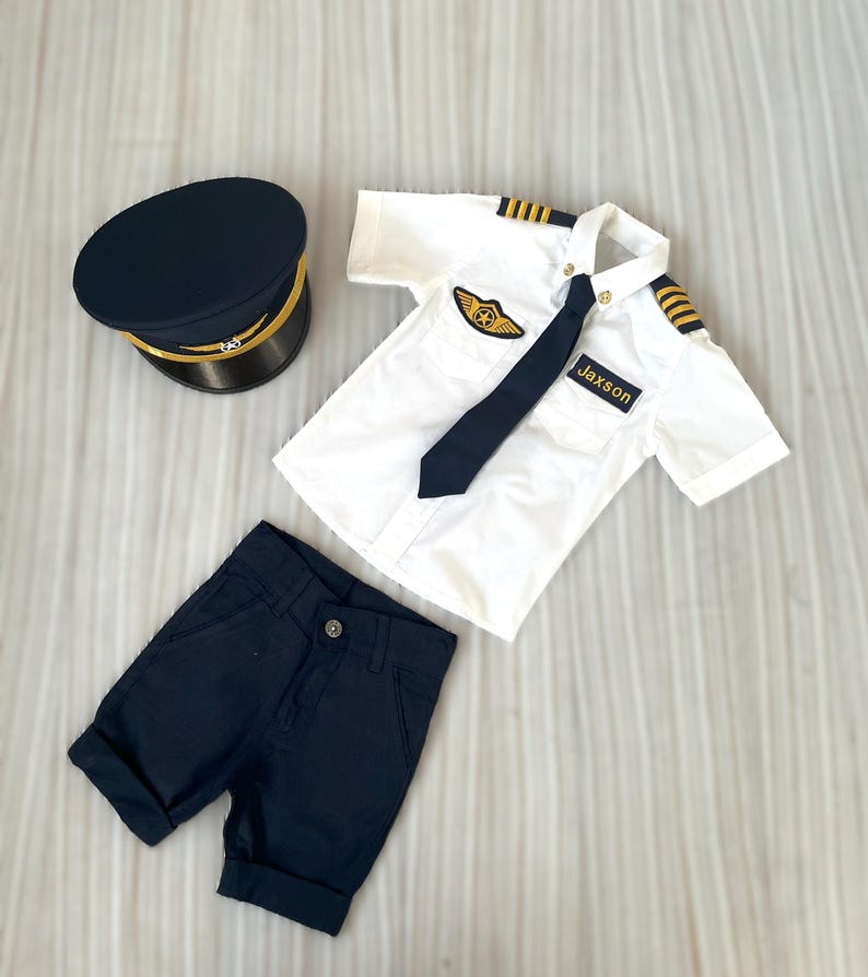 May include: A navy blue pilot's uniform set, including a hat, shirt, shorts, and tie. The white shirt has gold stripes on the shoulders and a patch with a gold emblem. The name "Jaxson" is on the shirt.